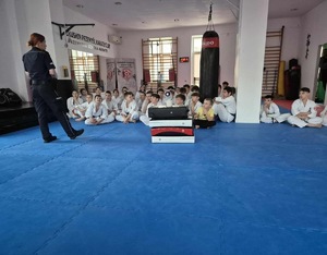 spotkanie policjantów z dziećmi ze szkoły karate w Przemyślu. uczniowie ubrani w kimona siedzą na niebieskich matach. policjantka wygłasza prelekcję.