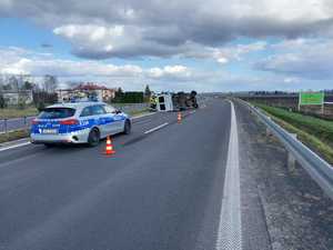 Zdarzenie drogowe w m. Hurko. Na zdjęciu widoczny jest oznakowany radiowóz, a w oddali na drodze , leży przewrócony na lewy bok biały bus . przy busie stoją strażacy.