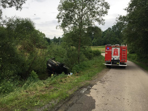 samochód przewrócony do góry kołami w zaroślach , w przydrożnym potoku, w tle radiowóz strażacki