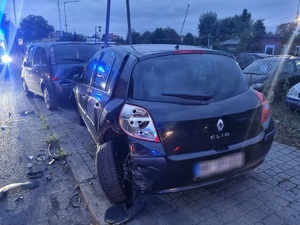 zdjęcie przedstawia samochód marki Renault w kolorze ciemnym, stojący na chodniku. pojazd ma uszkodzony tylną , lewa stronę tj. nadkole i wykrzywione koło. za pojazdem stoją kolejne samochody na chodniku