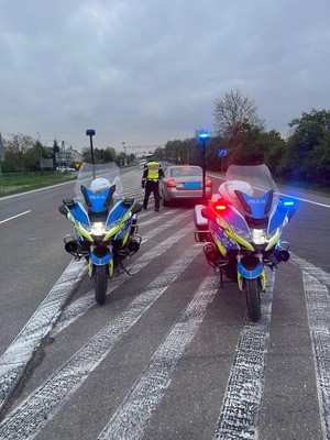 Zdjęcie kolorowe przedstawia dwa motocykle policyjne stojące na jezdni a przed nimi widoczny policjant podchodzący do zatrzymanego pojazdu.