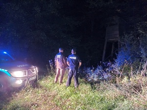 zdjęcie wykonane nocą. policjant i funkcjonariusz straży leśnej stoją przed amboną leśną . Obok stoi radiowóz policyjny.