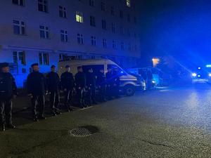 policjanci oddają hołd zmarłemu ratownikowi medycznemu w Siedlicach. Przed komendą stoją Radiowozy z włączonymi światłami błyskowymi w kolorze niebieskim i obok policjanci.