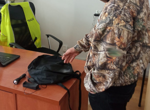 mężczyzna wskazujący dłonią na plecak i latarkę położone na biurku. w tle krzesło i kamizelka odblaskowa z napisem policja
